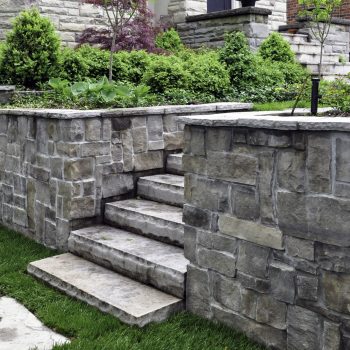 Luxury landscaping shows natural stone steps, a retaining wall with coping, and oversized flagstone stepping stones.